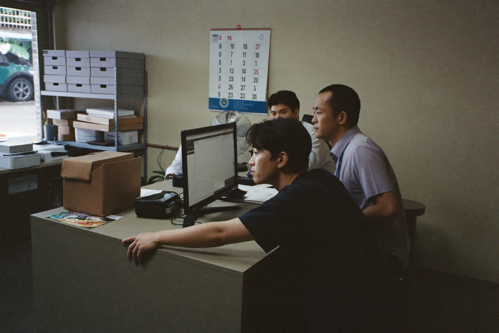 Three colleagues at one monitor in a cramped back office, all looking at the screen rather than the camera. Box files on steel shelving, a wall calendar and a desk fan behind them.