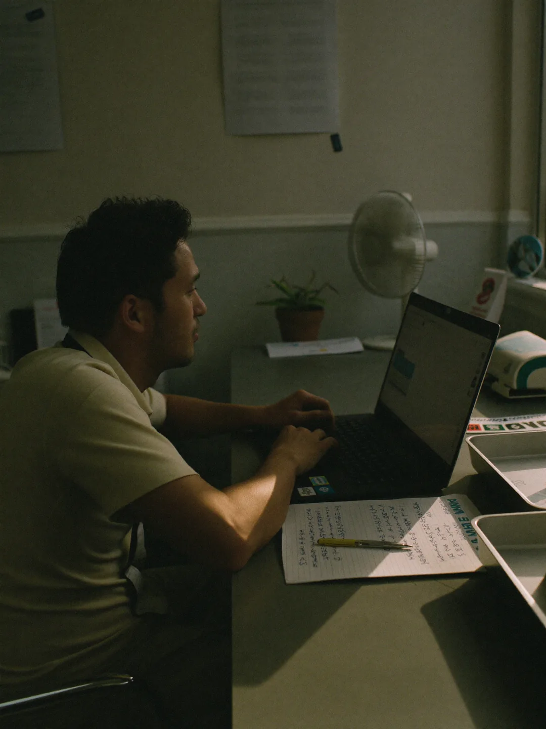 One person at a laptop in late afternoon light, reading, with an open notebook of handwritten notes and a pen on the desk beside the keyboard.