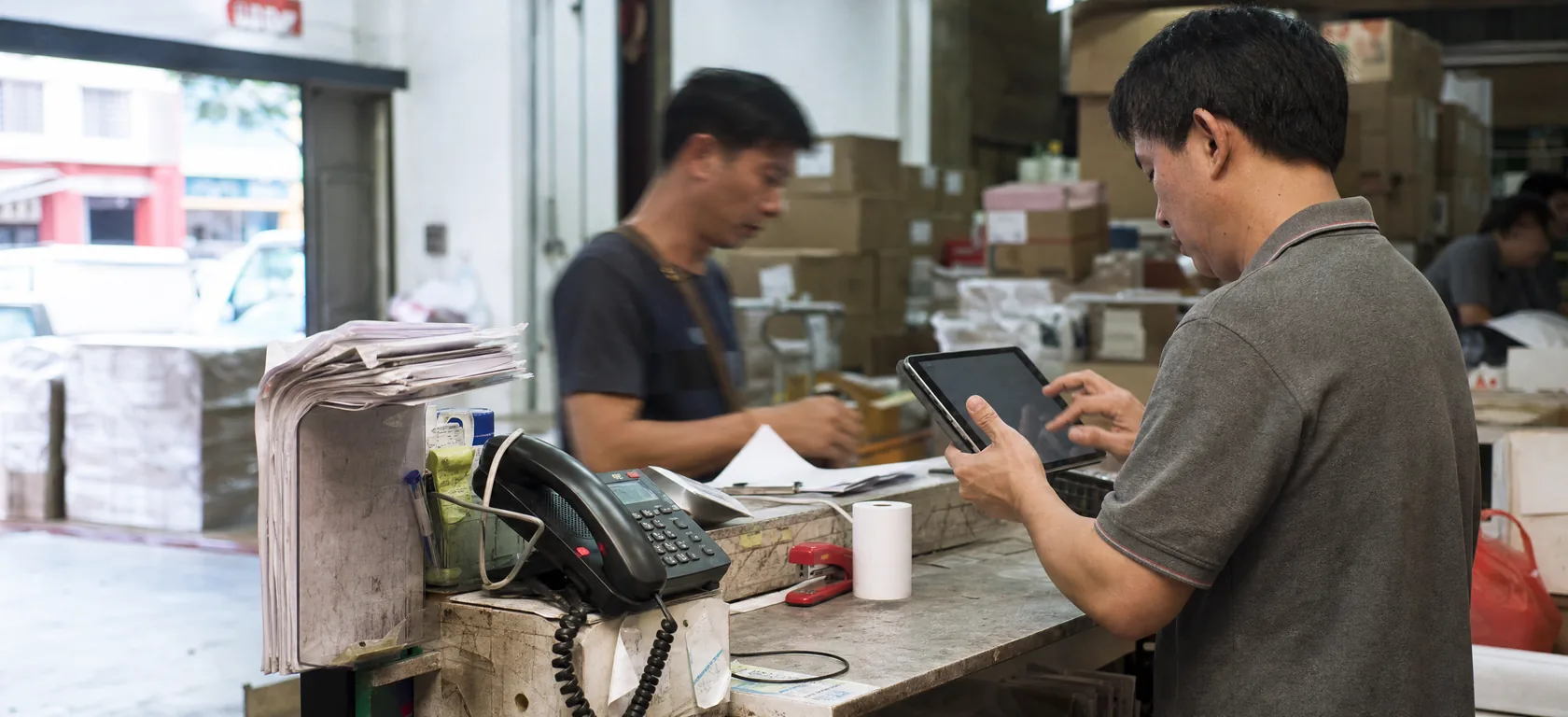 A man at a goods counter entering something on a tablet held in one hand, a spiked stack of paper dockets and a desk phone on the counter beside him, a colleague working behind and cartons stacked on racking. The street is visible through the open shutter.