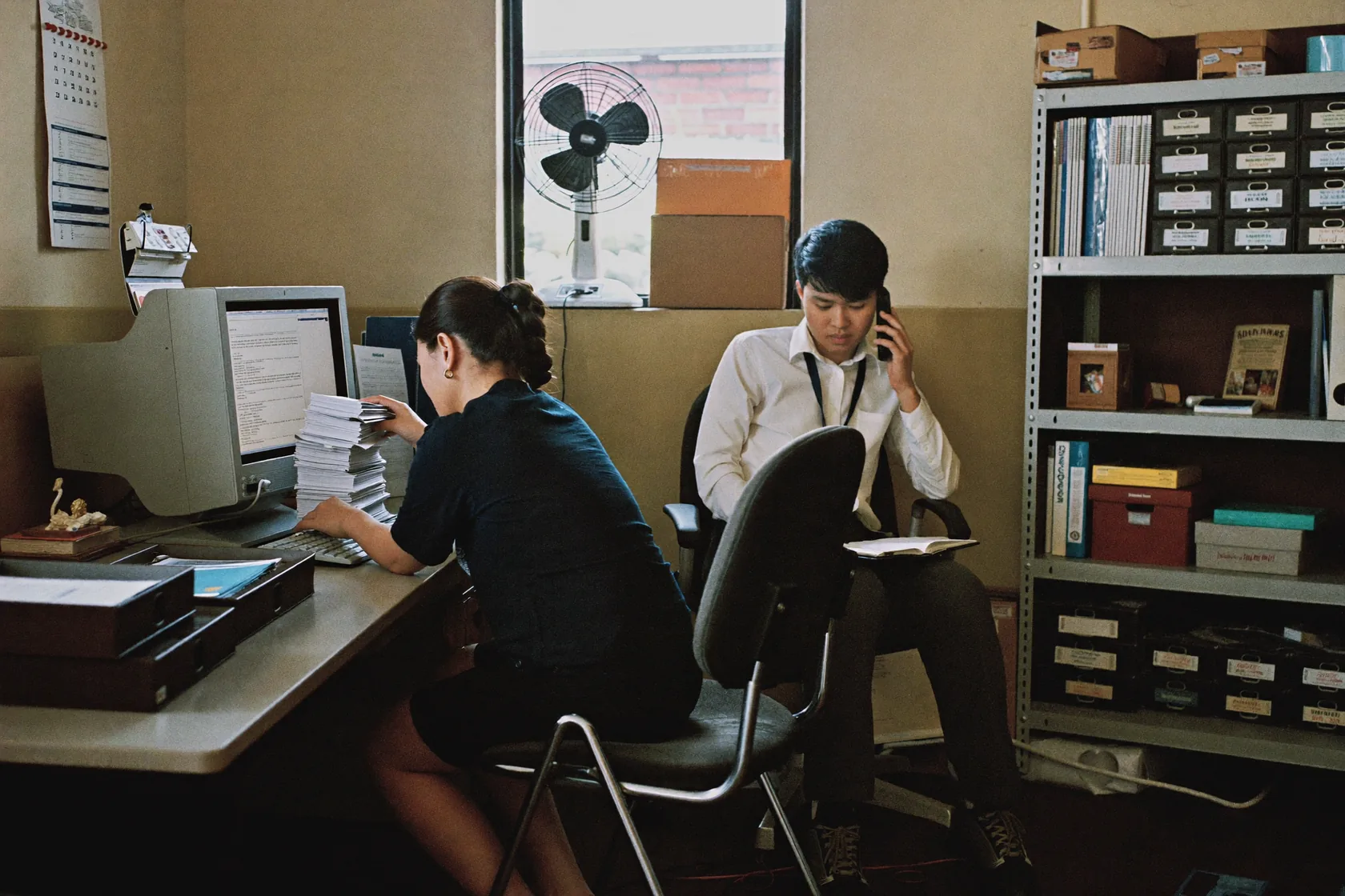 An administrator keying a thick stack of paper dockets into an old desktop system, a colleague at the next chair with an open notebook. Filing shelves, a wall calendar and a window fan behind them.