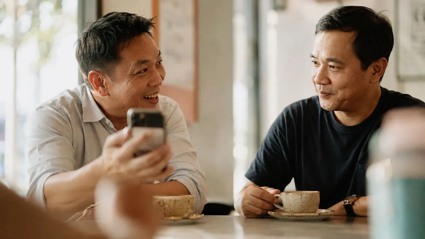 Two men at a cafe table; one leans forward grinning and turns his phone towards the other, who smiles back but has visibly started thinking about something else.